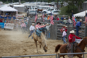 Zwei Reiter auf Pferden bei einem Wettbewerb namens Charreada, umgeben von Menschen, Bannern und Fahnen, Zeltplanen, geparkten Fahrzeugen und Bäumen im Hintergrund.