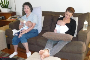 Zwei Frauen sitzen auf einem Sofa und halten Babys, mit Kissen auf dem Sofa, einer weißen Wand im Hintergrund und einem Schreibtisch mit Büchern und einer Zimmerpflanze links daneben.