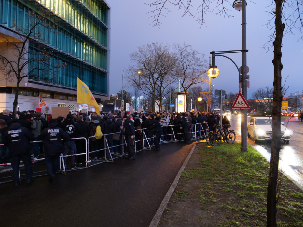 Eine große Gruppe von Menschen mit Protestschildern steht vor einem Gebäude in Berlin, mit Barrikaden, Fahrrädern, Schildern, Bäumen und Gras.