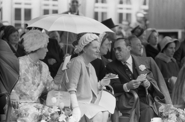 Schwarzes und weißes Bild von Queen Elizabeth II und Prince Philip, die mit Schirmen auf Stühlen sitzen, umgeben von Menschen mit Hüten und Büchern, mit einem Gebäude und Blumenarrangements im Hintergrund.