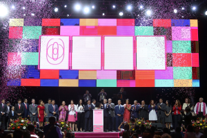 Eine Gruppe von Menschen steht auf einer Bühne mit einem Rednerpult, Blumengestecken auf beiden Seiten und einem großen Bildschirm im Hintergrund während der Eröffnungszeremonie des 2018 China International Film Festival.