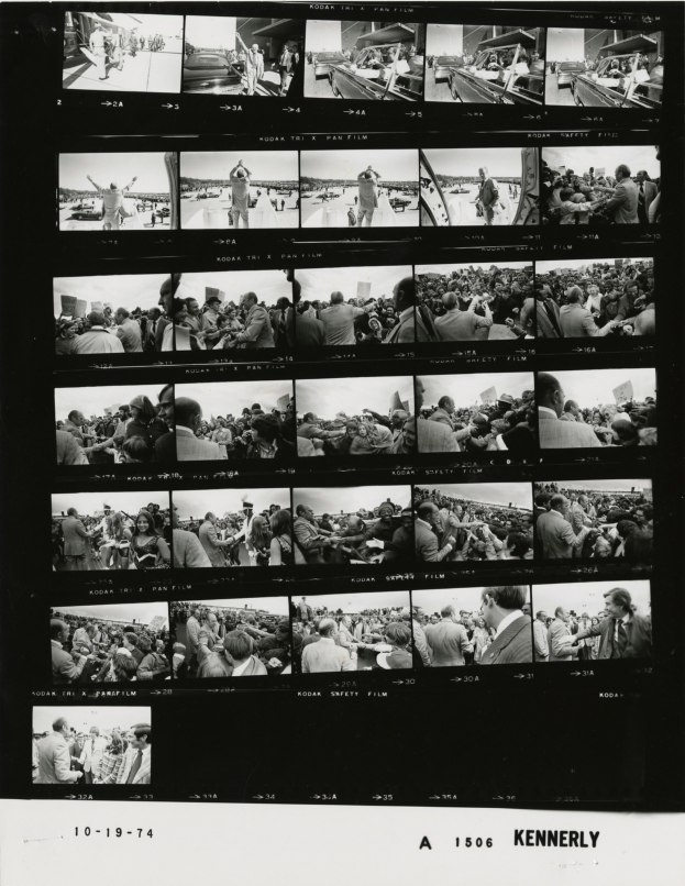 Schwarzes und weißes Foto einer vielfältigen Menge, die fröhlich in die Kamera schaut, während sie auf einen Fernseher schaut.