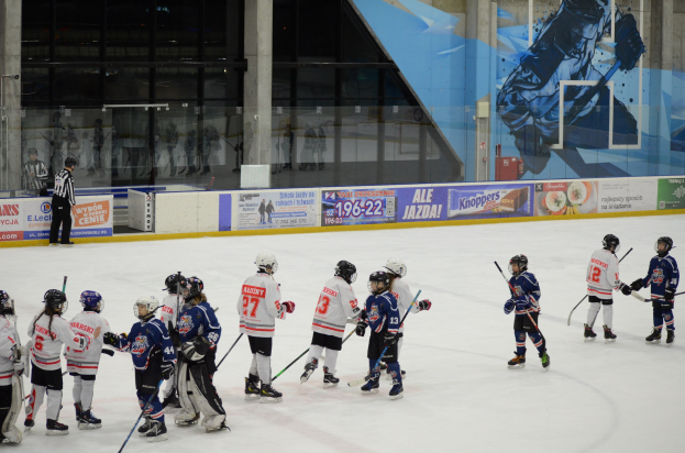 Eine Gruppe junger Menschen in Helmen und Hockey-Ausrüstung, die Eisockey auf einer Indoor-Eisbahn spielen, mit Bannern, Säulen und Glaswänden im Hintergrund.