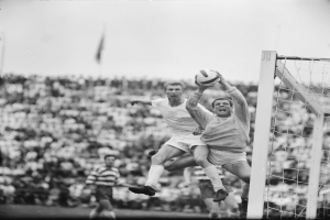 Zwei Männer spielen Fußball vor einem Publikum, mit einem Torpfosten auf der rechten Seite und einer Fahne im Hintergrund, in Schwarz-Weiß.
