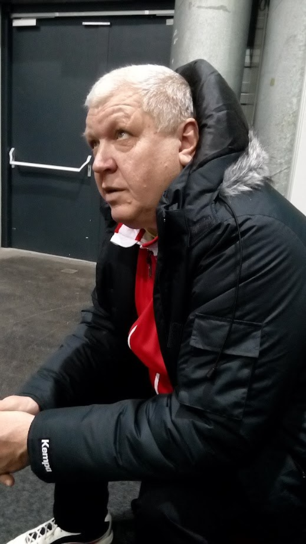 Ein Mann in schwarzer Jacke und rotem Hemd sitzt vor einer Tür mit Griff, einer Säule und einer Wand auf dem Boden und scheint tief in Gedanken versunken.