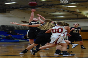 Mehrere Frauen in Trikots auf einem Basketballfeld mit einem Basketball und Netz sowie Zuschauern im Hintergrund unter Deckenbeleuchtung.