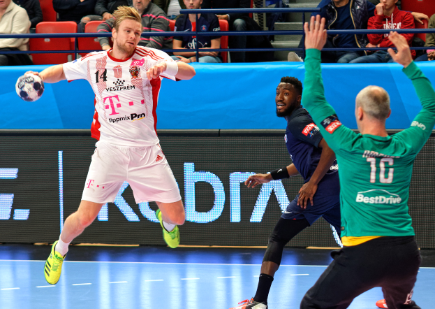 Eine Gruppe von Männern spielt Handball auf einem Platz mit einem Ball in der Mitte, während Zuschauer von Stühlen und Geländern in einer Arena zusehen, mit einem Schild im Hintergrund, auf dem 'Futsal-Weltmeisterschaft 2019 - Bayern München gegen Paris Saint-Germain' steht.