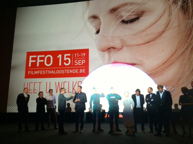 Eine Gruppe von Menschen auf einer Bühne mit einem Mann, der ein Mikrofon hält, in der Mitte, ein Display hinter ihnen mit dem Text "Foto Filmfestivaloostende 2015" und einem Bild einer Frau und einigen sitzenden Personen neben dem Bildschirm.