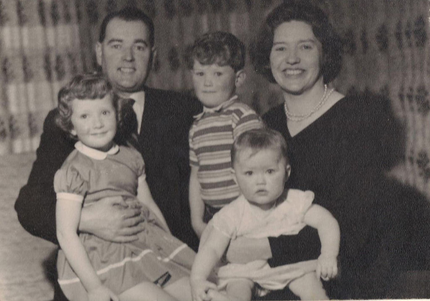 Eine Familie mit fünf Personen, darunter ein Mann in Anzug und Krawatte links, eine Frau rechts und drei strahlende Kinder, die gemeinsam posen.