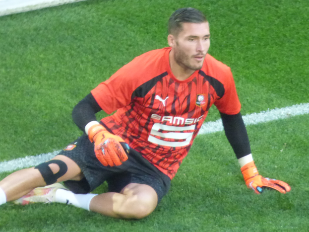 Ein Fußballspieler sitzt auf dem Boden, trägt ein rotes T-Shirt, schwarze Shorts, weiße Socken und orangefarbene Baseball-Handschuhe.