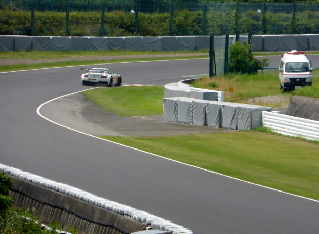 Ein Rennwagen fährt eine Rennstrecke entlang, im Hintergrund ein Polizeiwagen, umgeben von Gras, Pflanzen, Bäumen und Zaun, mit einem Fahrzeug auf der rechten Seite und Text unten.