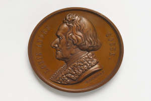 Eine runde Bronzetafel mit einem Porträt von Louis Andre Goss und einer Inschrift, vor einem weißen Hintergrund.