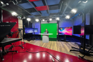 Ein Mann sitzt an einem Schreibtisch in einem TV-Studio mit einem Laptop und anderen Gegenständen, umgeben von Kamerastativen und Equipment, mit Bildschirmen und Deckenleuchten im Hintergrund.