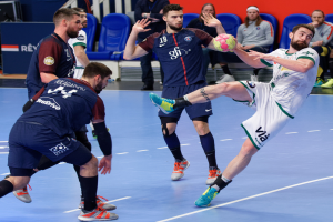 Eine Gruppe von Männern, die Handball auf einem Platz spielen, wobei einer den Ball hält, und Zuschauern im Hintergrund.