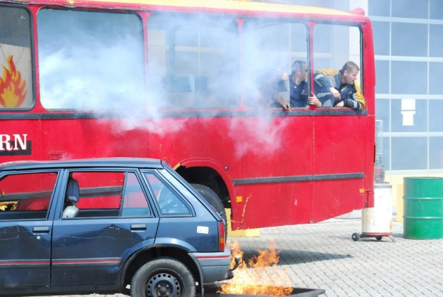 Ein roter Doppeldeckerbus mit Rauch, der aus ihm herauskommt, mit drei sichtbaren Passagieren, der neben einem Auto geparkt ist, vor einem Gebäude mit Glasfenstern und einem Fass auf der rechten Seite.