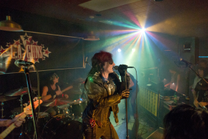 Eine Frau in einer schwarzen Jacke singt auf der Bühne in ein Mikrofon, während ein Mann mit Sonnenbrille und Stirnbinde ein Instrument spielt und ein "LA Guns"-Plakat sowie Diskoleuchten im Hintergrund zu sehen sind.