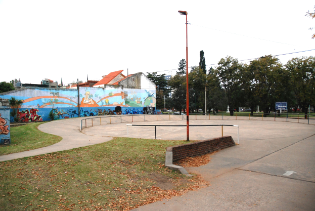 Skate-Anlage mit Wandgemälde an einem Gebäude, umgeben von Gras, trockenen Blättern, Laternen, Geländern und einer bemalten Wand, vor einem Hintergrund aus Bäumen, Gebäuden, Fahrzeugen und Himmel.