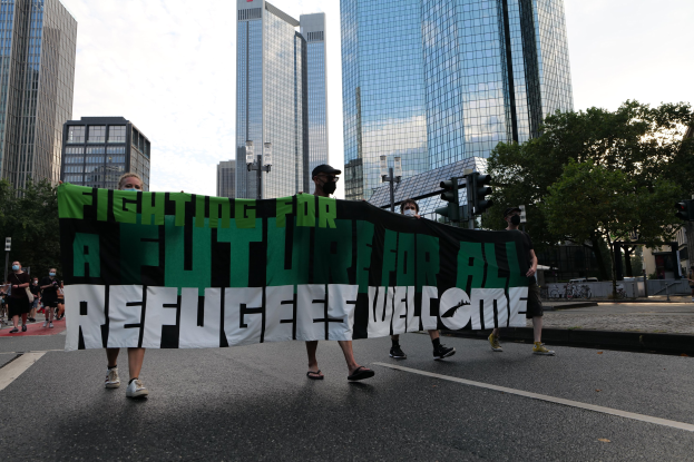 Eine Gruppe von Menschen geht eine Straße entlang und hält ein Banner mit der Aufschrift "Kämpfen für eine Zukunft für alle - Willkommen Flüchtlinge" mit Bäumen und Gebäuden am Straßenrand und Verkehrsampeln im Hintergrund unter einem bewölkten Himmel.