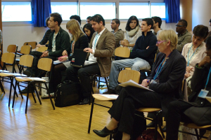 Mehrere Personen sitzen auf Stühlen in einem Raum, tragen ID-Karten und halten einen Laptop und einen Stift, umgeben von leeren Stühlen, Fenstern mit Vorhängen und einer Tasche auf dem Boden.