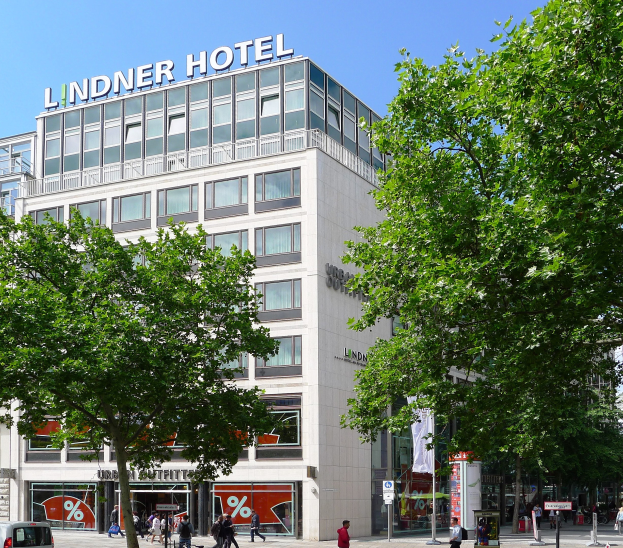 Großes gläsernes Gebäude mit der Aufschrift "Lindner Hotel" in Berlin, Deutschland, mit Bäumen, Fahrzeugen, Passanten und Himmel im Hintergrund.