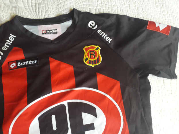 Ein schwarzes und rotes Fussballtrikot mit einer roten und schwarzen Streifen, einem Logo und Text, das auf einer weissen Oberfläche liegt.