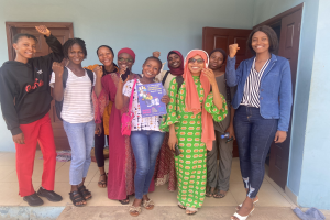 Eine Gruppe von Frauen vor einem Schulgebäude in Nigeria, die strahlend Bücher in den Händen halten, mit einem Tuch auf dem Boden und Türen im Hintergrund.