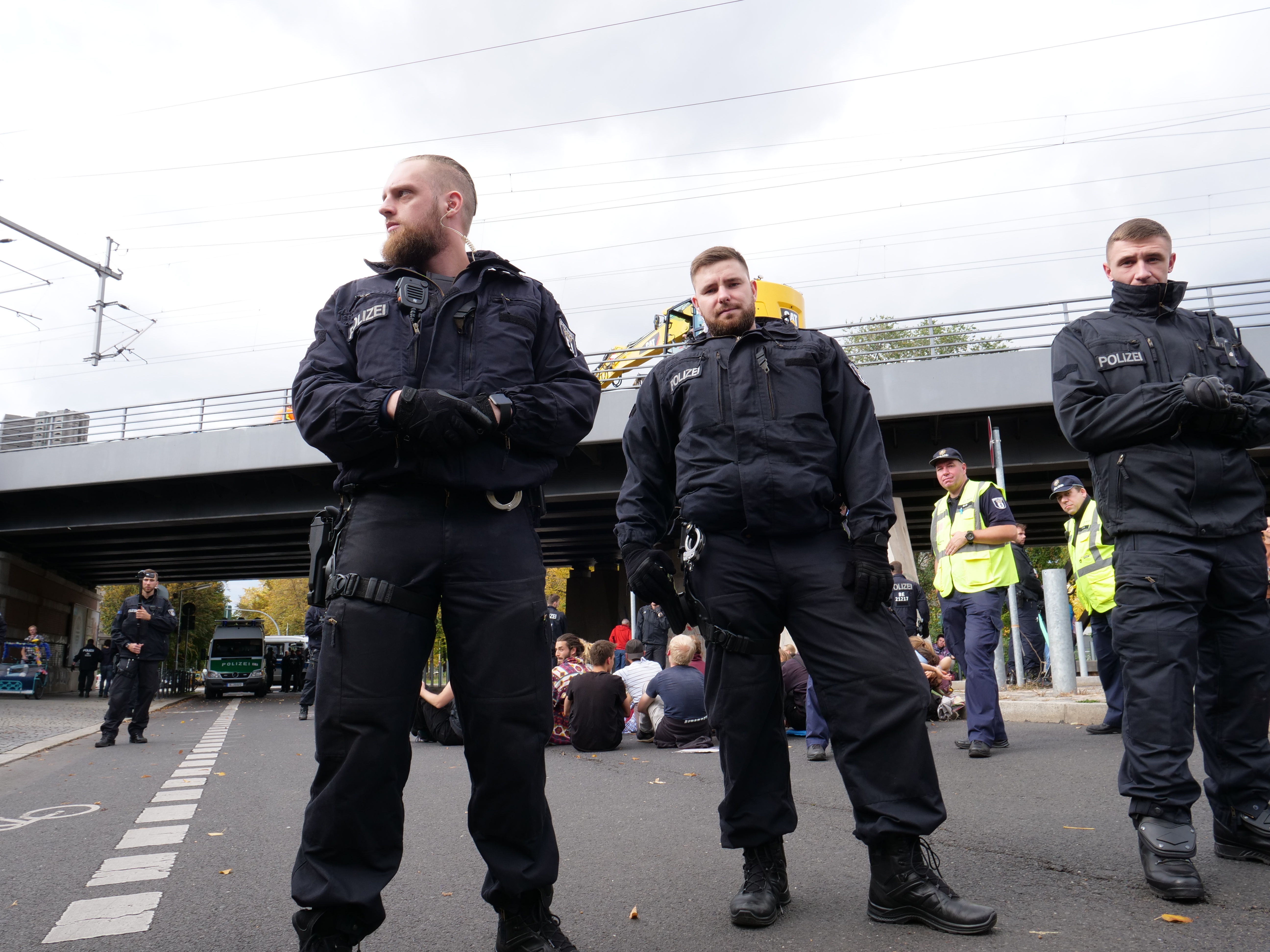Eine Gruppe von Polizisten steht an der Straßenseite mit Menschen, Fahrzeugen, einer Brücke, Bäumen, Mästen, Drähten und einem klaren blauen Himmel im Hintergrund.