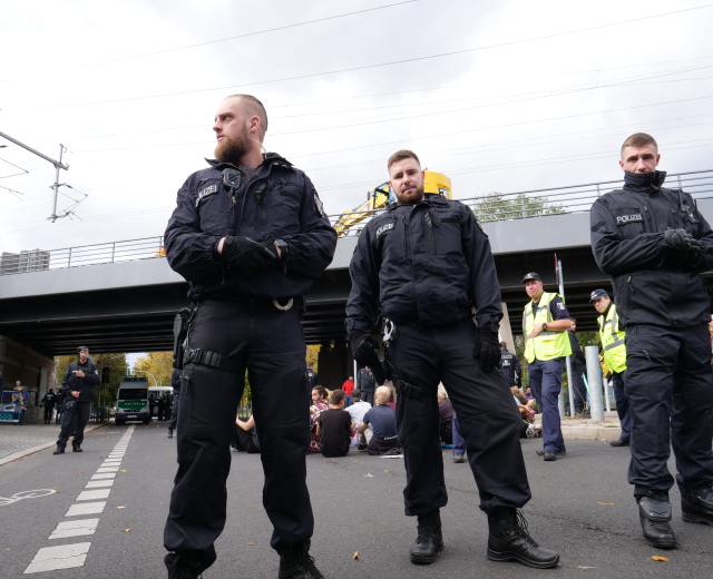 Eine Gruppe von Polizisten steht an der Straßenseite mit Menschen, Fahrzeugen, einer Brücke, Bäumen, Mästen, Drähten und einem klaren blauen Himmel im Hintergrund.