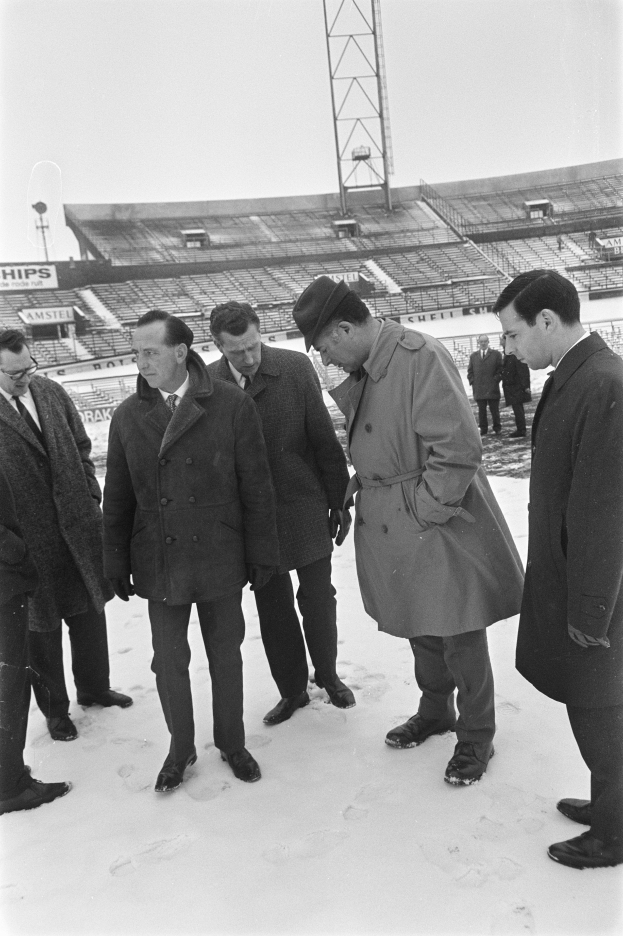 Eine Gruppe von Männern in Mänteln und Schuhen steht zusammen im Schnee, mit einem Stadion und einem Turm im Hintergrund unter einem klaren Himmel.