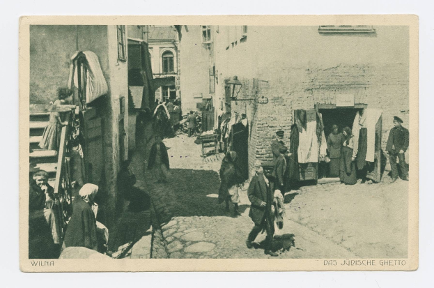 Schwarz-weißes Foto von Menschen, die eine Straße im jüdischen Ghetto von Wilna entlanggehen, mit Gebäuden im Hintergrund und Text unten.