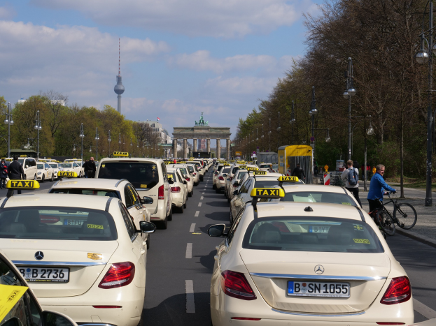 Eine lange Reihe von Taxis, die entlang einer belebten Straße in Berlin, Deutschland, geparkt sind, mit Fahrradfahrern und Fußgängern auf dem Gehweg, flankiert von Laternen und Bäumen und Gebäuden, einem Bogen und einem Turm im Hintergrund unter einem bewölkten Himmel.