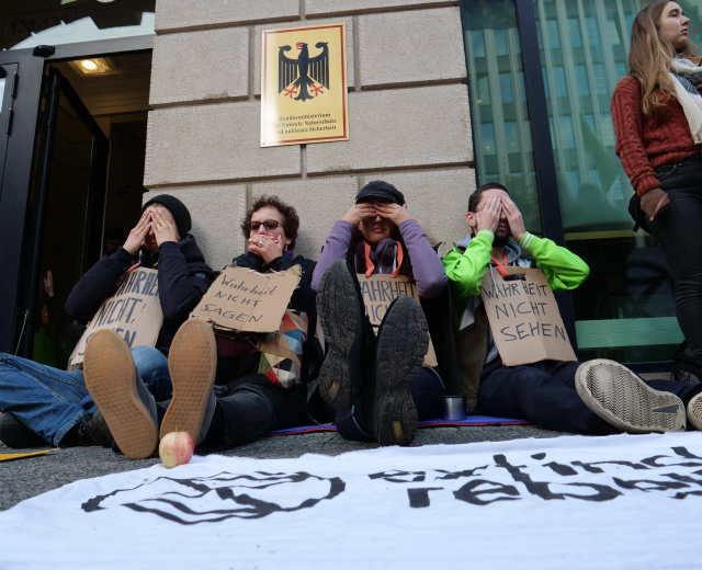Eine Gruppe von Menschen sitzt vor einem Gebäude auf dem Boden und hält Schilder, mit einer Fahne auf der Straße und einer Frau rechts daneben, die besorgt aussieht.