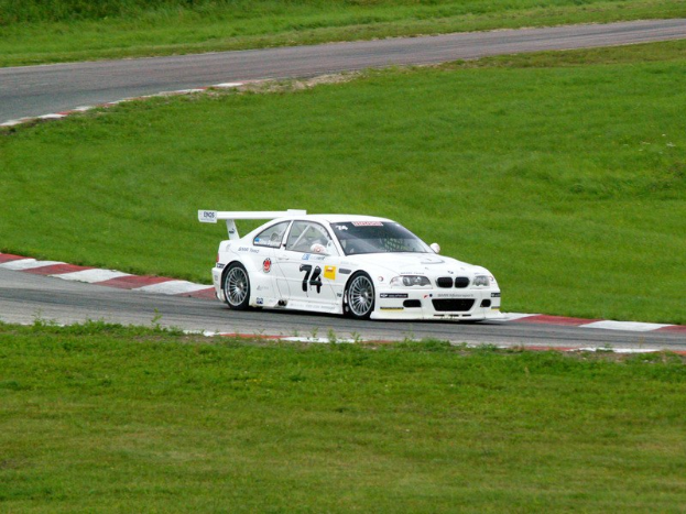 Weißer BMW M3 GT3 Rennwagen fährt auf einer Rennstrecke mit grünem Gras und Pflanzen im Hintergrund.