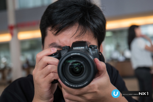 Ein Mann in einem schwarzen T-Shirt hält eine Kamera vor sein Gesicht, während eine Frau in einem weißen Kleid in der rechten Ecke steht.