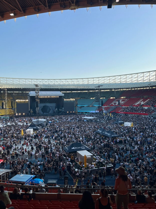 Eine große Menschenmenge, die in einer Arena sitzt und steht, umgeben von Gittern, Zelten und Lichtern, mit einem Dach und weiteren Lichtern oben, was auf ein Konzert in der O2 Arena in London hindeutet.