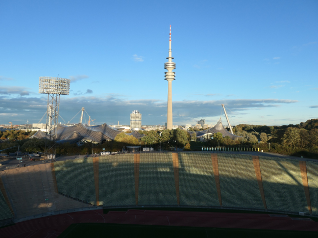 Außenansicht des Olympiastadions in Berlin, Deutschland, mit dem Fernsehturm (Fernsehturm) im Hintergrund, umgeben von Bäumen, Gebäuden und beleuchteten Bereichen unter einem bewölkten Himmel.