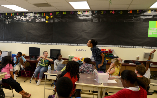 Ein Klassenzimmer mit Kindern an Tischen mit verschiedenen Gegenständen, einer Lehrerin in der Mitte und einer Tafel im Hintergrund.