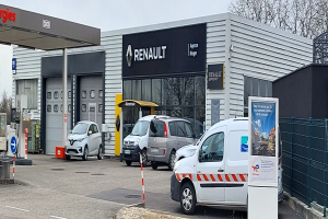 Ein Renault-Autohaus mit parkenden Autos davor, umgeben von einem Metallzaun, einem Schild, Pfosten, einer Baumgruppe und einem bewölkten Himmel.