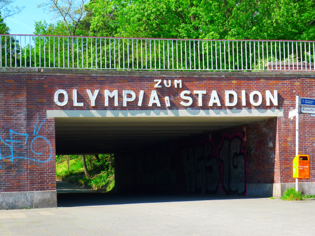 Der Eingang zum Olympiastadion in Berlin, Deutschland, mit einer Brücke mit Text, einem Metallzaun, einer Schautafel, einem Kasten, Pflanzen, Gras, einer Baumgruppe und einem bewölkten Himmel.