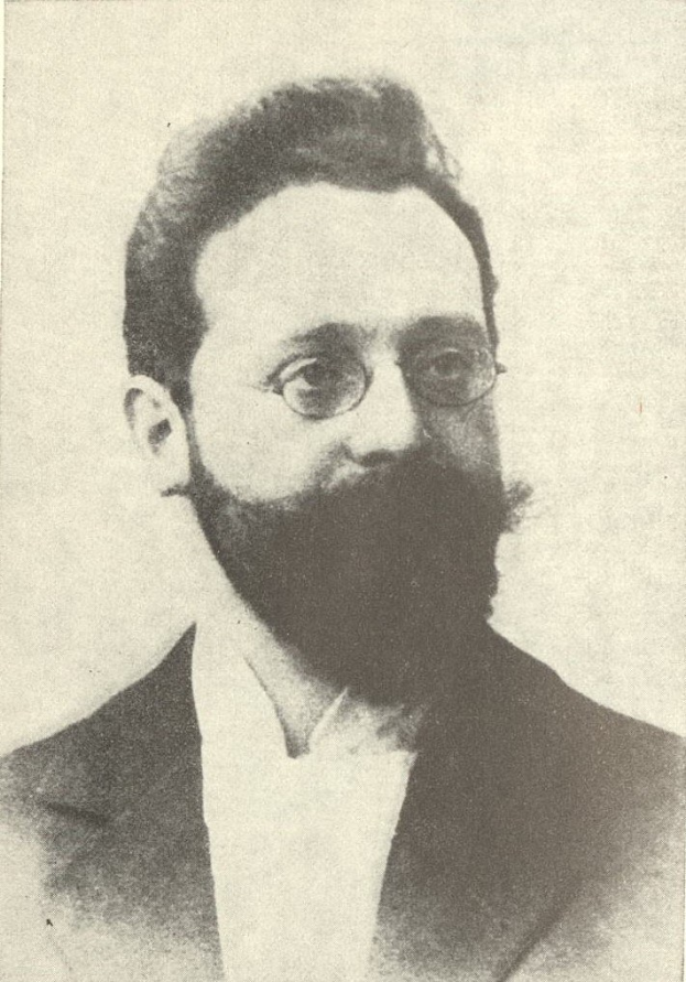 Schwarzes und weißes Porträt eines bärtigen Mannes in Anzug und Krawatte mit Brille, der ernst in die Ferne schaut.