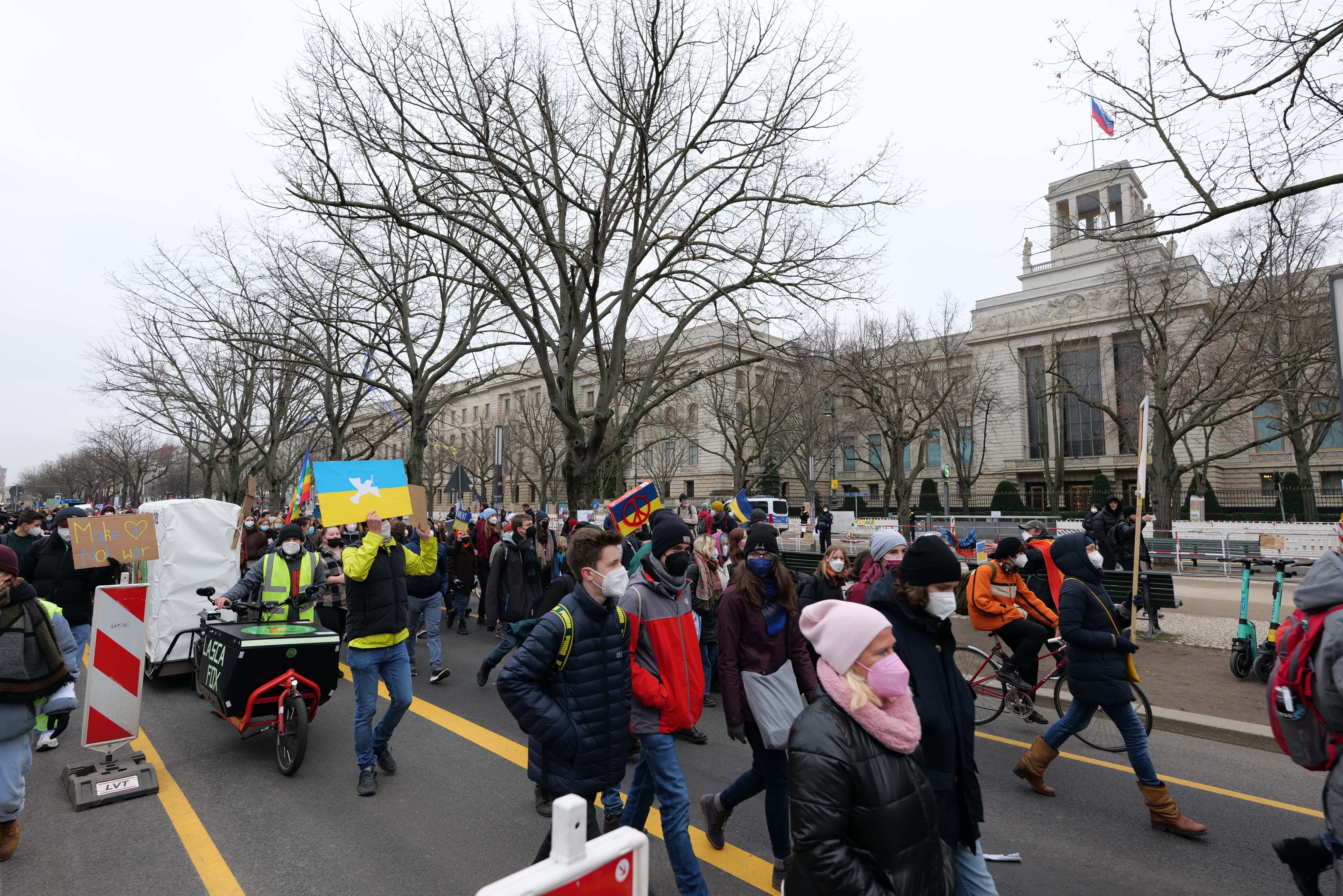Eine große Protestmarsch mit Menschen, die eine Straße in Washington, D.C. entlanggehen, einige halten Schilder und andere fahren Fahrr√°der, mit Bäumen und einem klaren blauen Himmel im Hintergrund.