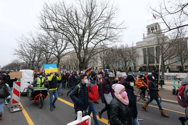 Eine große Protestmarsch mit Menschen, die eine Straße in Washington, D.C. entlanggehen, einige halten Schilder und andere fahren Fahrr√°der, mit Bäumen und einem klaren blauen Himmel im Hintergrund.