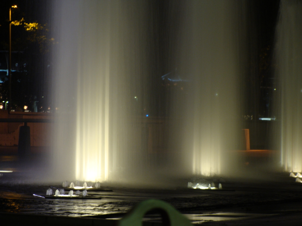 Ein Wasserfall im Vordergrund mit Bäumen, beleuchteten Polen und Gebäuden im Hintergrund.