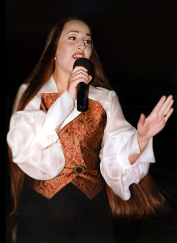 Frau in weißem Hemd und brauner Weste, die selbstbewusst in ein Mikrofon singt und es festhält, während ihr Haar zu einem Dutt zurückgebunden ist.