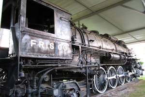 Ein Zug ist in der Mitte auf Schienen mit Gras darunter und einer Decke mit Pfählen darüber zu sehen.