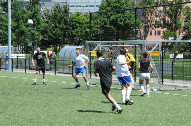 Personen spielen Fußball auf einem Feld mit einem Zaun und einem Tor in der Mitte, einem Pfosten und einem Ball oben links und Bäumen und Gebäuden im Hintergrund.