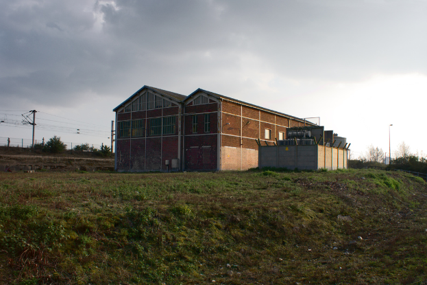 Eine Szene mit Schuppen, Transformatoren, elektrischen Polen mit Kabeln, Zäunen, einer Straßenlaterne, Bäumen und einem bewölkten Himmel.
