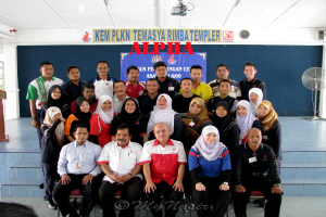 Eine Menschenmenge, die vor einer Bühne strahlt und posiert, mit einer Tafel mit Text an der Wand dahinter, einem Tischventilator rechts, einem Podium links und einem Projektor oben im Bild.