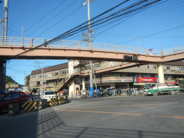 Eine Brücke überspannt eine vielbefahrene Straße mit Fahrzeugen, Strommästen mit Drähten, Gebäuden, Bäumen und einem klaren blauen Himmel im Hintergrund.