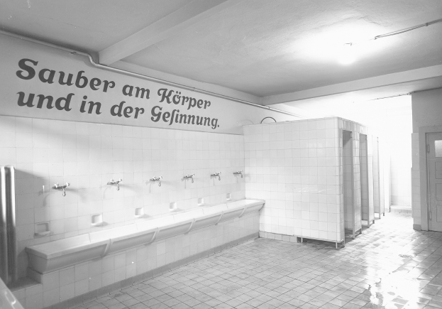 Ein Schwarz-Weiß-Foto einer öffentlichen Toilette mit einem gefliesten Boden, einer textbedeckten Wand, ein paar Armaturen, einer Tür und einer Deckenlampe.